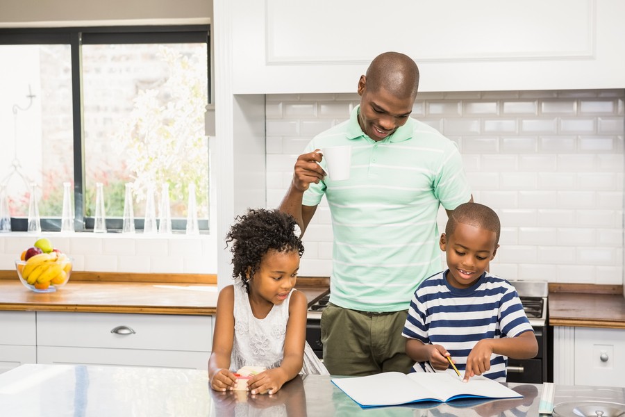  Image is of a family with one parent and two children gathered in the kitchen looking at homework.