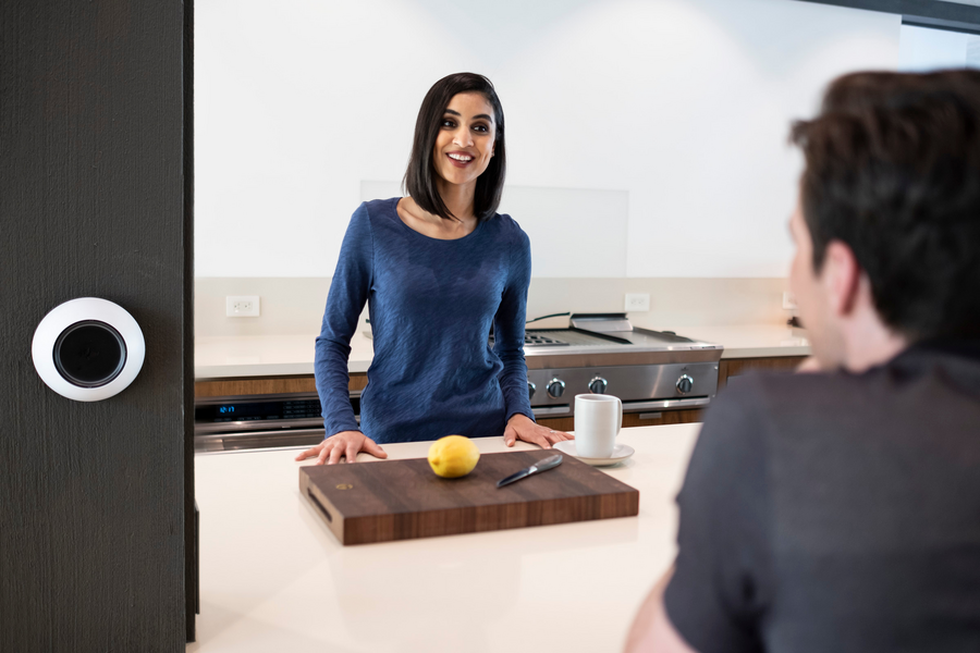 Two people have a conversation in a kitchen with a Josh.ai device on the wall in the foreground.